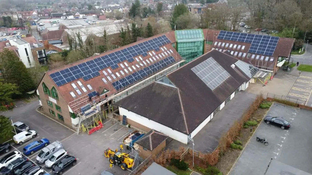 Aerial drone photograph of a large commercial solar panel installation in Dorset by Up Top Solar, powered by Up Top Roofing. Extensive solar PV array on pitched clay tile roof with professional scaffolding and qualified roofing team on site installing solar panels. Full building aerial view showing scale of commercial solar project.